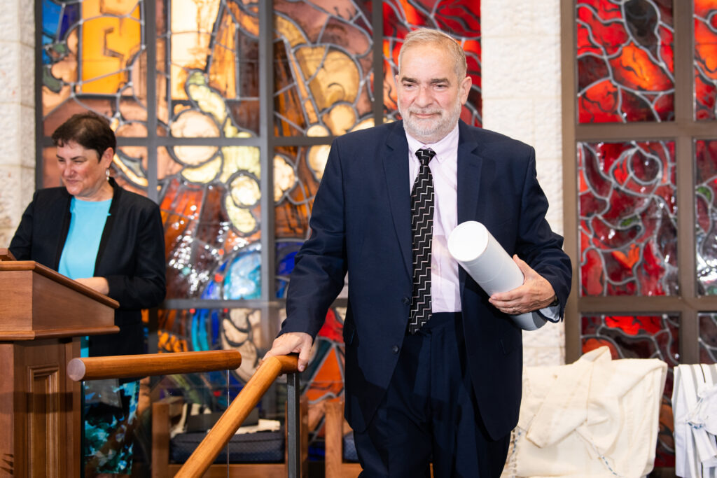 Rabbi Jacob Staub leaves the bimah at RRC's graduation ceremony, Doctor of Human Letters award in hand. Rabbi Deborah Waxman stands at the podium, looking down. 